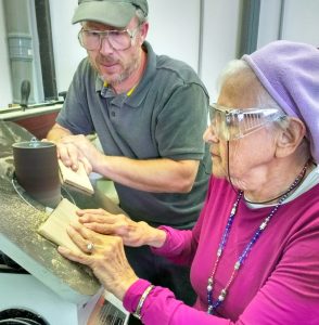 A ninety-year-old woman smooths a piece of wood on the oscillating spindle sander.